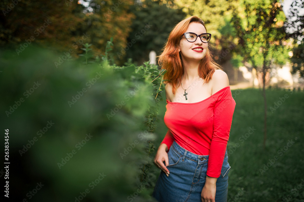Attractive redhaired woman in eyeglasses, wear on red blouse and jeans skirt posing at green park. Attractive redhaired woman in eyeglasses, wear on red blouse and jeans skirt posing at green park.