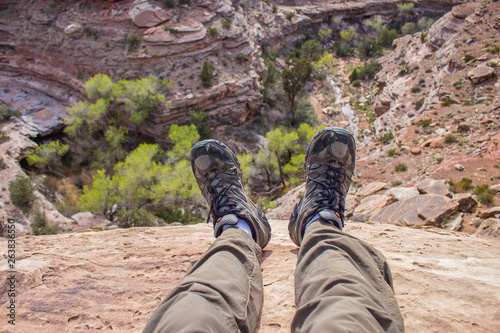 A hiker dangles his feet over the edge of a cliff looking down into a desert canyon in this first person pov photo. 