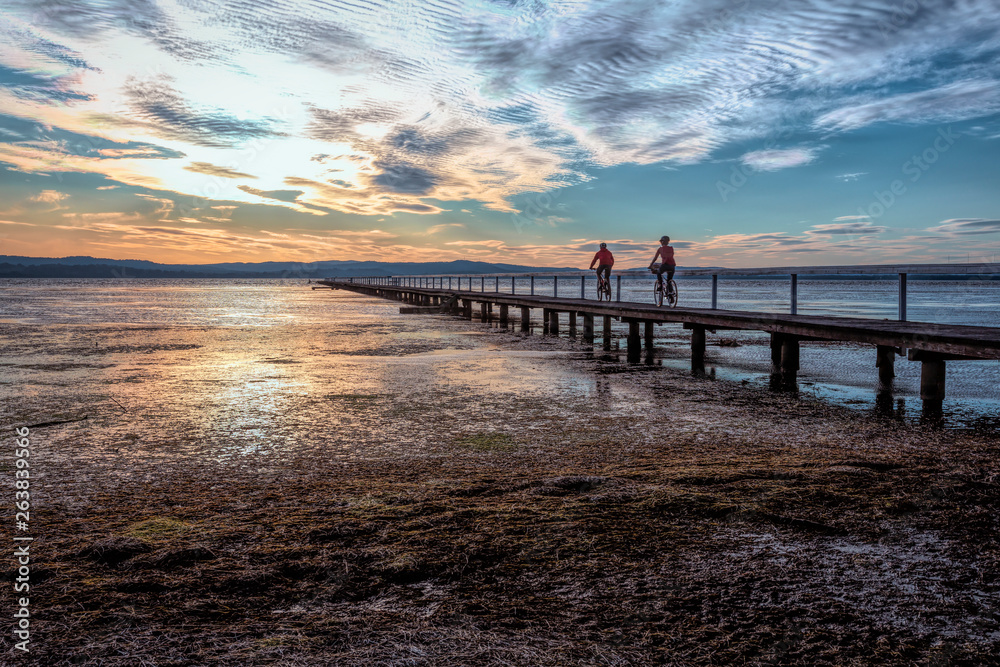 Fototapeta premium Rowerzyści na długim molo na dużym jeziorze pływowym o zachodzie słońca.