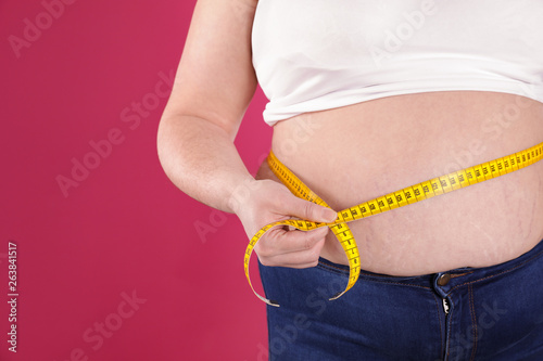 Overweight woman measuring waist with tape on color background, closeup. Space for text