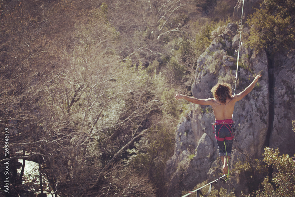 Obraz premium A man is walking along a stretched sling. Highline in the mountains. Man catches balance. Performance of a tightrope walker in nature. Highliner on the background of valley.