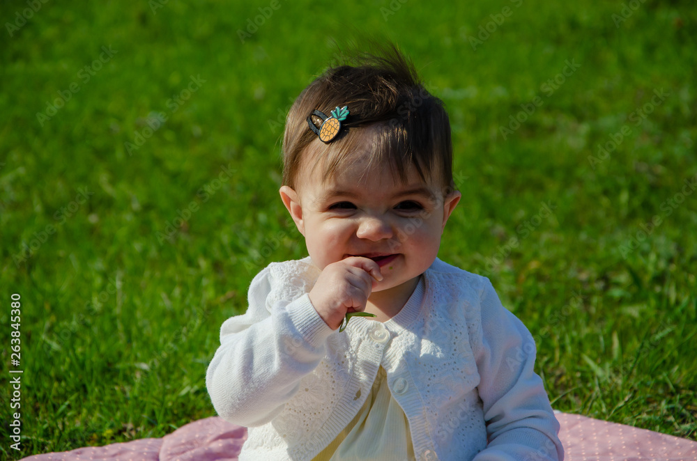 Fototapeta premium Baby in bright clothes on a pink plaid on green grass in the park. Newborn baby outside on a warm day. Little girl plays on a plaid on a green lawn in spring