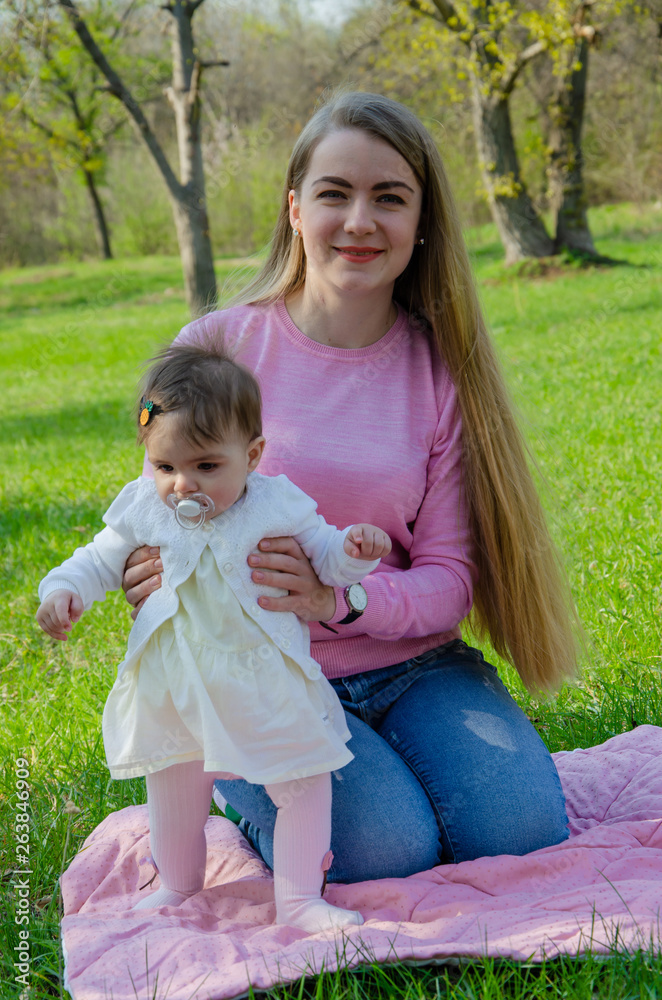 Fototapeta premium Mom with baby in bright clothes on a pink plaid on the green right. Family resting in the park on a warm day. Mom and little girl 10 months walk in the park