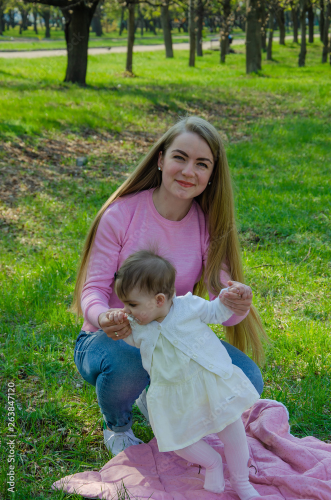 Mom with baby in bright clothes on a pink plaid on the green right. Family resting in the park on a warm day. Mom and little girl 10 months walk in the park Mom with baby in bright clothes on a pink plaid on the green right. Family resting in the park on a warm day. Mom and little girl 10 months walk in the park