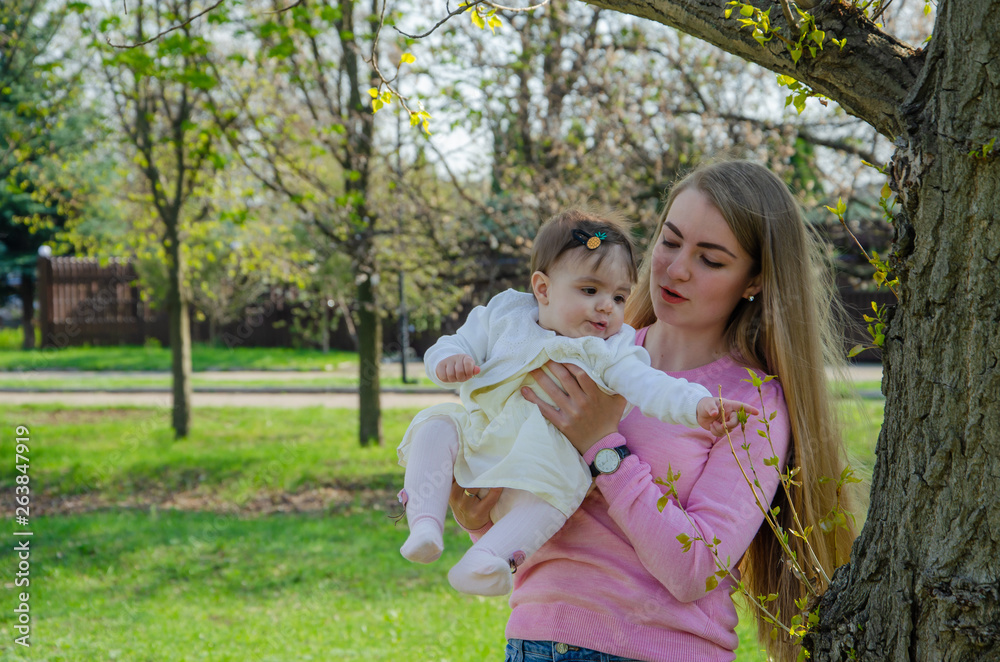 Mom with baby in bright clothes on a pink plaid on the green right. Family resting in the park on a warm day. Mom and little girl 10 months walk in the park