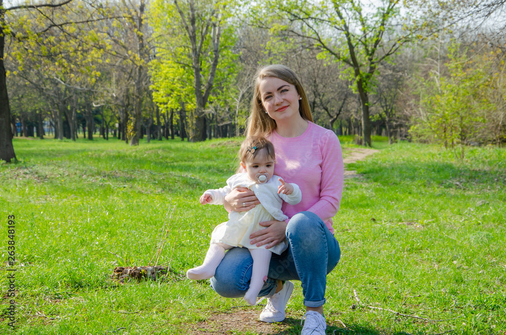 Fototapeta premium Mom with baby in bright clothes on a pink plaid on the green right. Family resting in the park on a warm day. Mom and little girl 10 months walk in the park