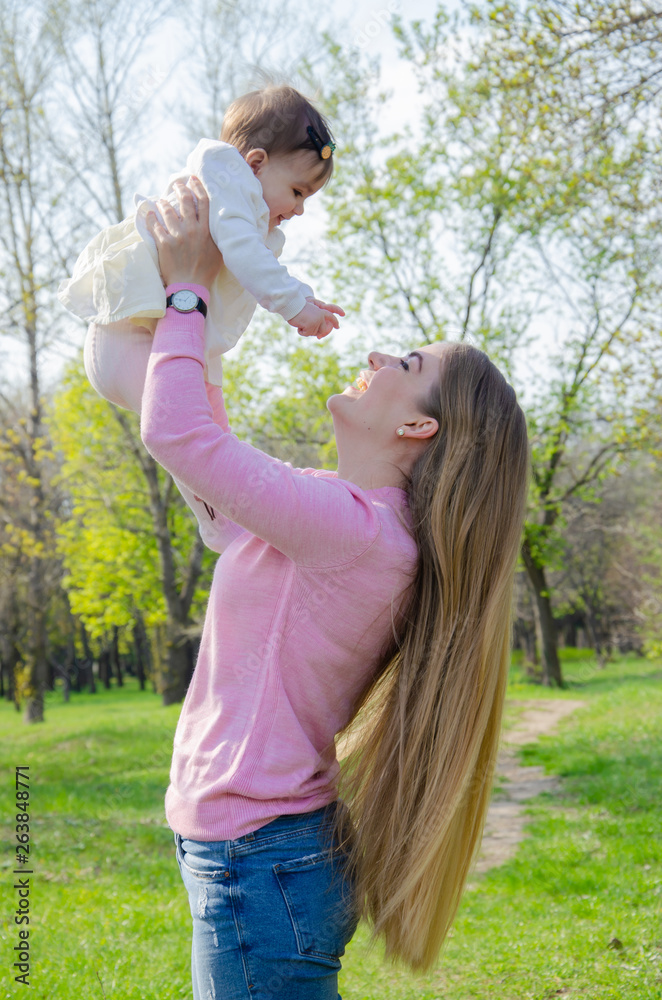 Obraz premium Mom with baby in bright clothes on a pink plaid on the green right. Family resting in the park on a warm day. Mom and little girl 10 months walk in the park