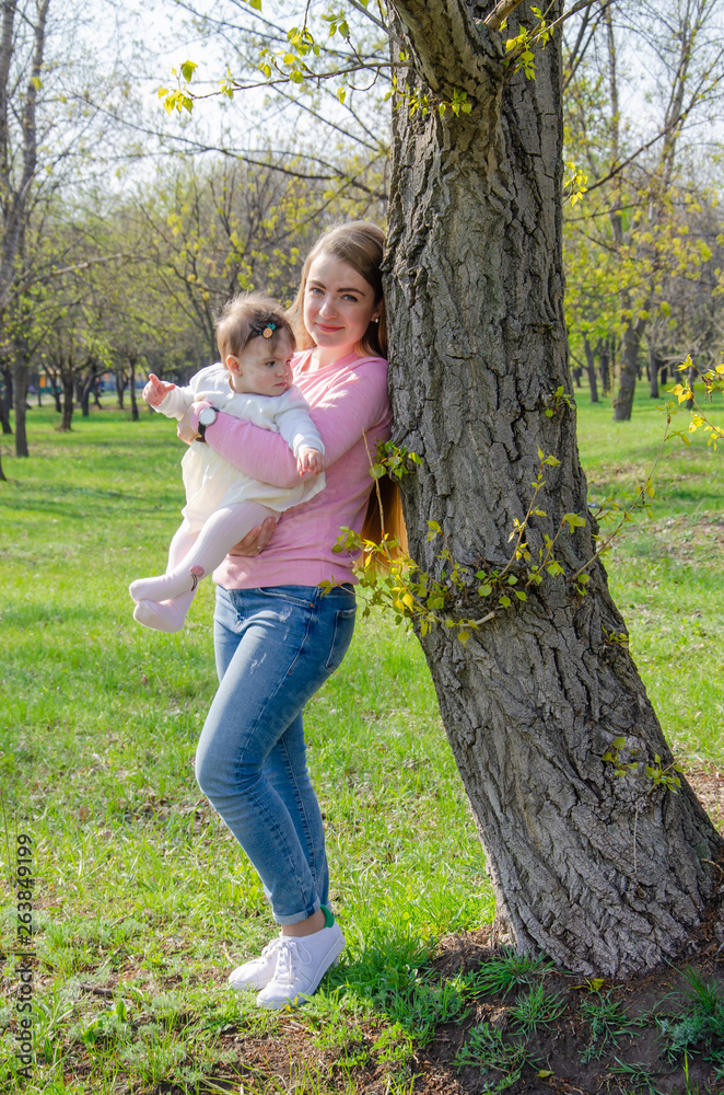 Fototapeta premium Mom with baby in bright clothes on a pink plaid on the green right. Family resting in the park on a warm day. Mom and little girl 10 months walk in the park