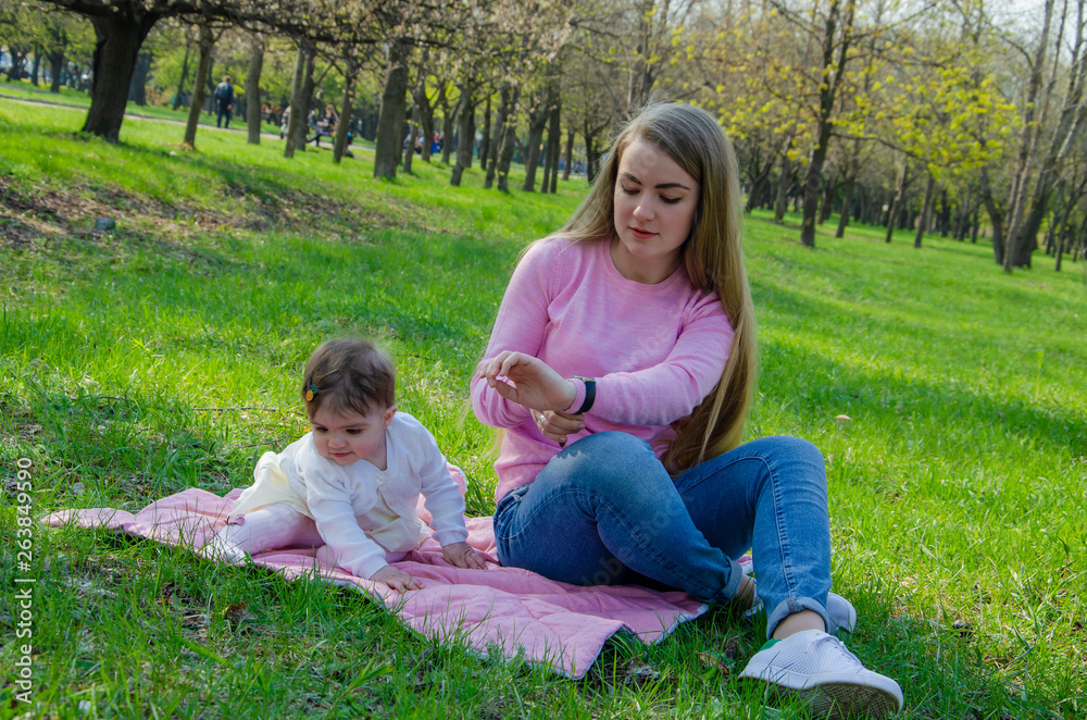 Mom with baby in bright clothes on a pink plaid on the green right. Family resting in the park on a warm day. Mom and little girl 10 months walk in the park Mom with baby in bright clothes on a pink plaid on the green right. Family resting in the park on a warm day. Mom and little girl 10 months walk in the park