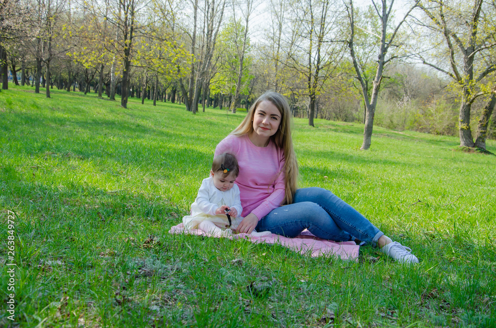 Mom with baby in bright clothes on a pink plaid on the green right. Family resting in the park on a warm day. Mom and little girl 10 months walk in the park Mom with baby in bright clothes on a pink plaid on the green right. Family resting in the park on a warm day. Mom and little girl 10 months walk in the park
