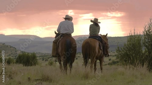 Cowboy and cowgirl stop for a rest sitting on horses, view of the mountain range and sunset ahead, 29 fps.