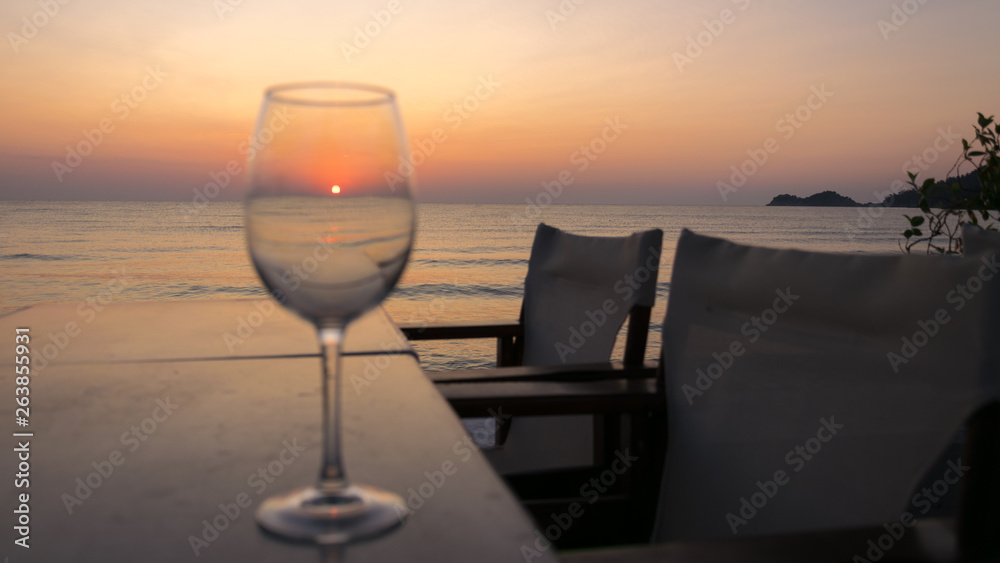 the sunrise seen through a glass on the table, with chairs and calm sea water.