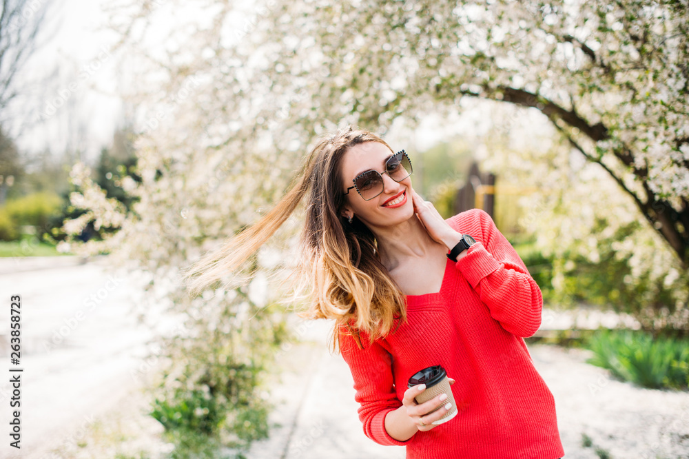 Beautiful happy young woman enjoying smell in a flowering spring garden. Female in sunglasses with long streaming hair and coffee to go dancing outdoor and smiling. Fashion Lifestyle People emotions Beautiful happy young woman enjoying smell in a flowering spring garden. Female in sunglasses with long streaming hair and coffee to go dancing outdoor and smiling. Fashion Lifestyle People emotions