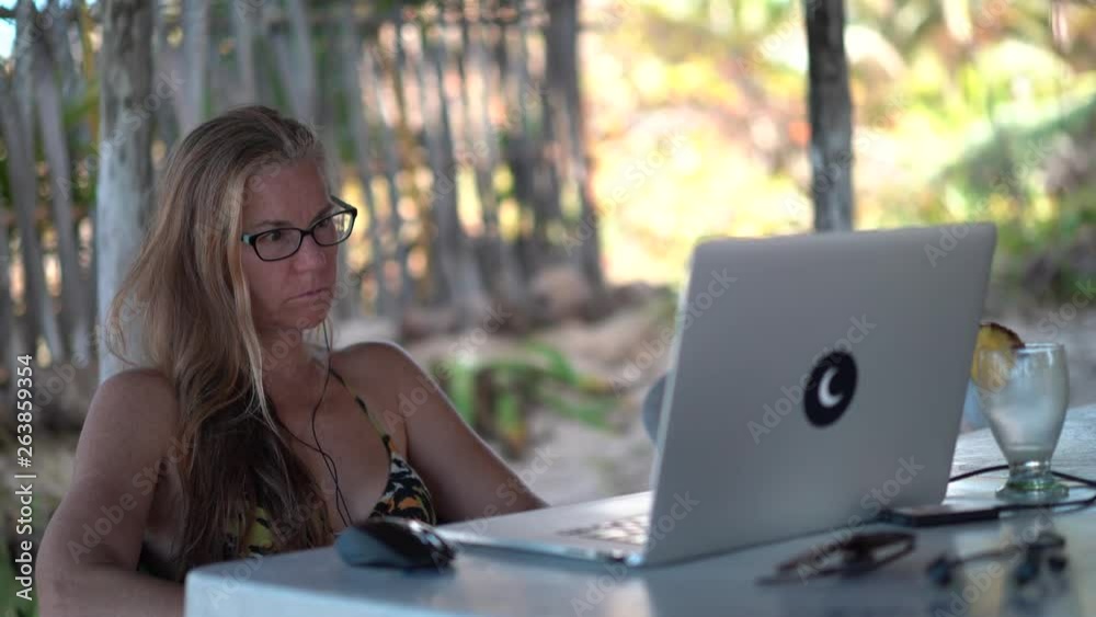 Middle aged blond woman in bikini with empty pina colada outside and working on a computer at a tropical beach resort. Middle aged blond woman in bikini with empty pina colada outside and working on a computer at a tropical beach resort.