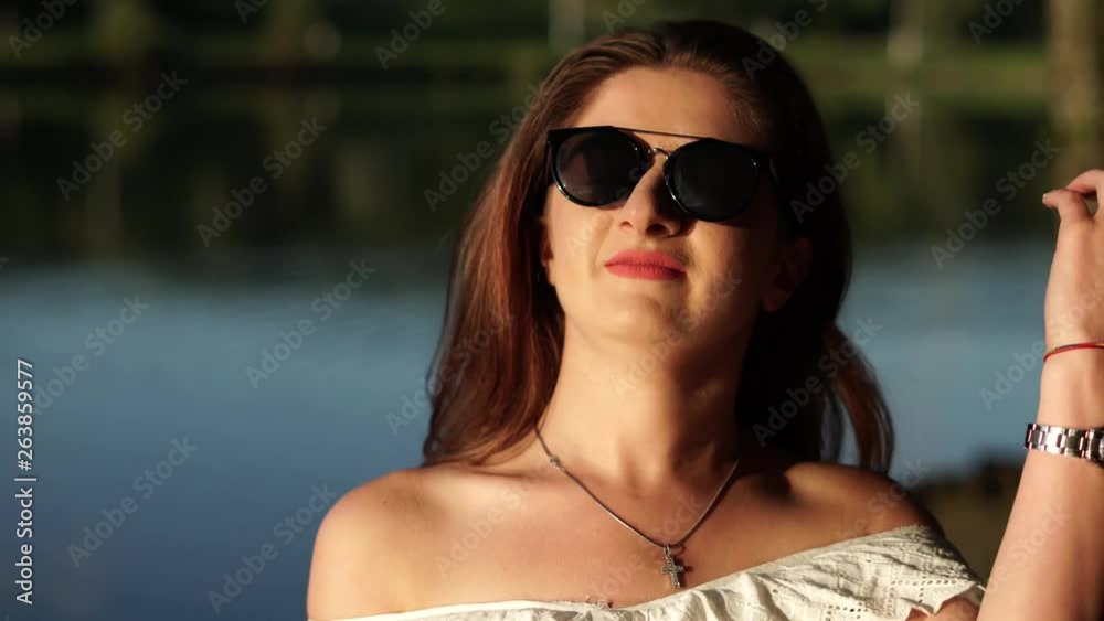 Close-up of attractive brunette in a white strapless dress and sunglasses gazing at nature and touching her hair on a bokeh lake background