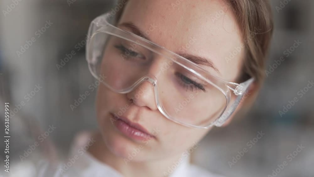 Close Up Portrait of Young Caucasian Female Scientist Wearing Protective Glasses in Chemical Laboratory Holding Flask With Gloves. Light Camera Movement Close Up Portrait of Young Caucasian Female Scientist Wearing Protective Glasses in Chemical Laboratory Holding Flask With Gloves. Light Camera Movement