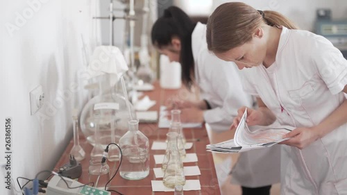 Female Caucasian Scientist in Bilogical Laboratory Checking Flasks With Transparent Liquid on Workbench and Compares Values On Paper Documents. Blurred Background photo