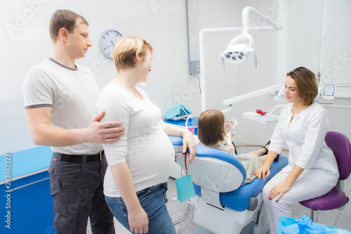 A female dentist communicates with a child and her family in a dental clinic. The mother of the girl is pregnant