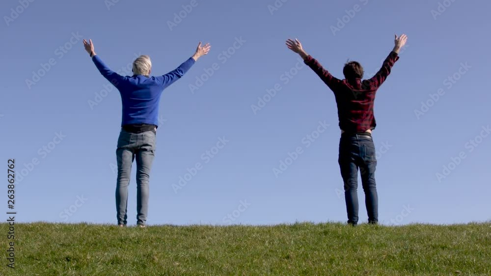 Two men standing up on green grass facing a blue background horizon raise their arms to the sky in yoga position and breathe at the same time.