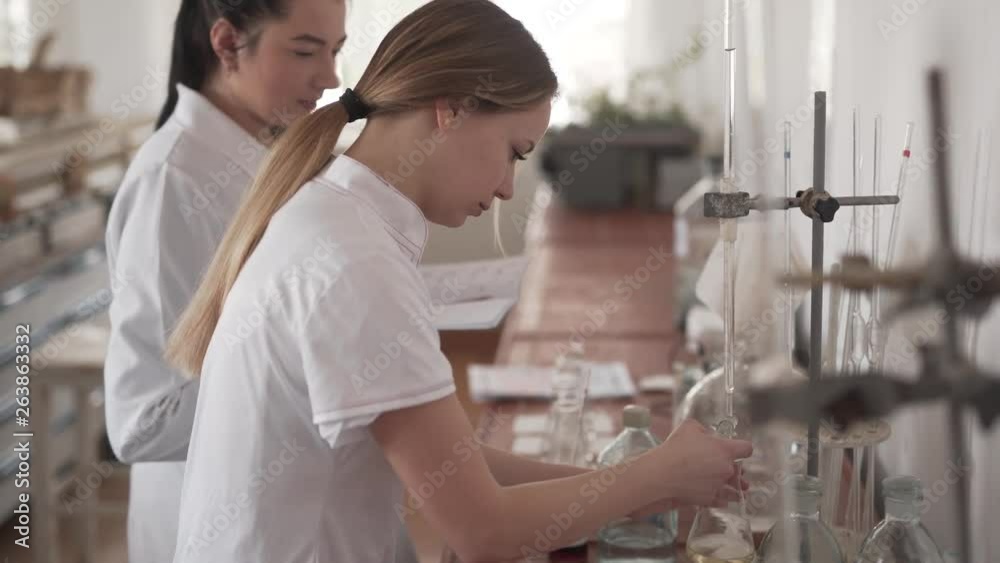 There are two female scientists in the laboratory. One takes notes and writes down the observations. The other conducts the experiment on pesticides. She uses the glass bulb with yellow liquid inside There are two female scientists in the laboratory. One takes notes and writes down the observations. The other conducts the experiment on pesticides. She uses the glass bulb with yellow liquid inside