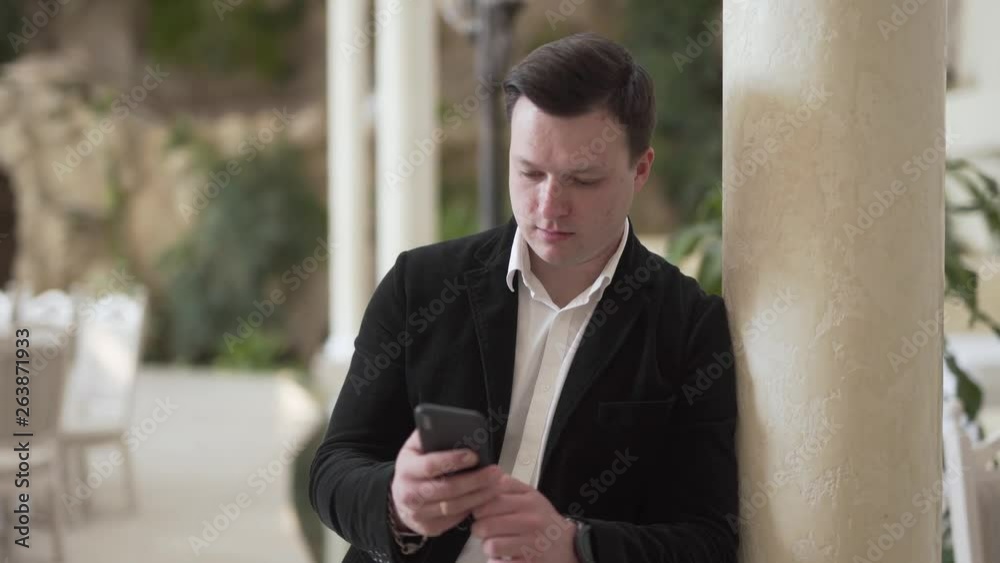 Young man in a black suit and white shirt is standing in a restauran, leaning on a column and clicking in the phone. Man is smiling. Young man in a black suit and white shirt is standing in a restauran, leaning on a column and clicking in the phone. Man is smiling.