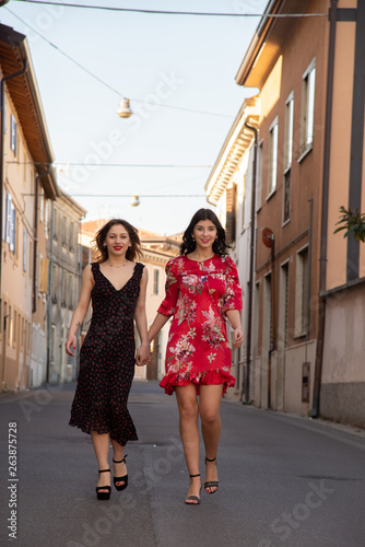 Full-length portrait of two friends, a blonde in a black dress and the other brunette in a red dress. Breathe a full sense and enjoy freedom. They walk in the middle of the street holding hands. Conce