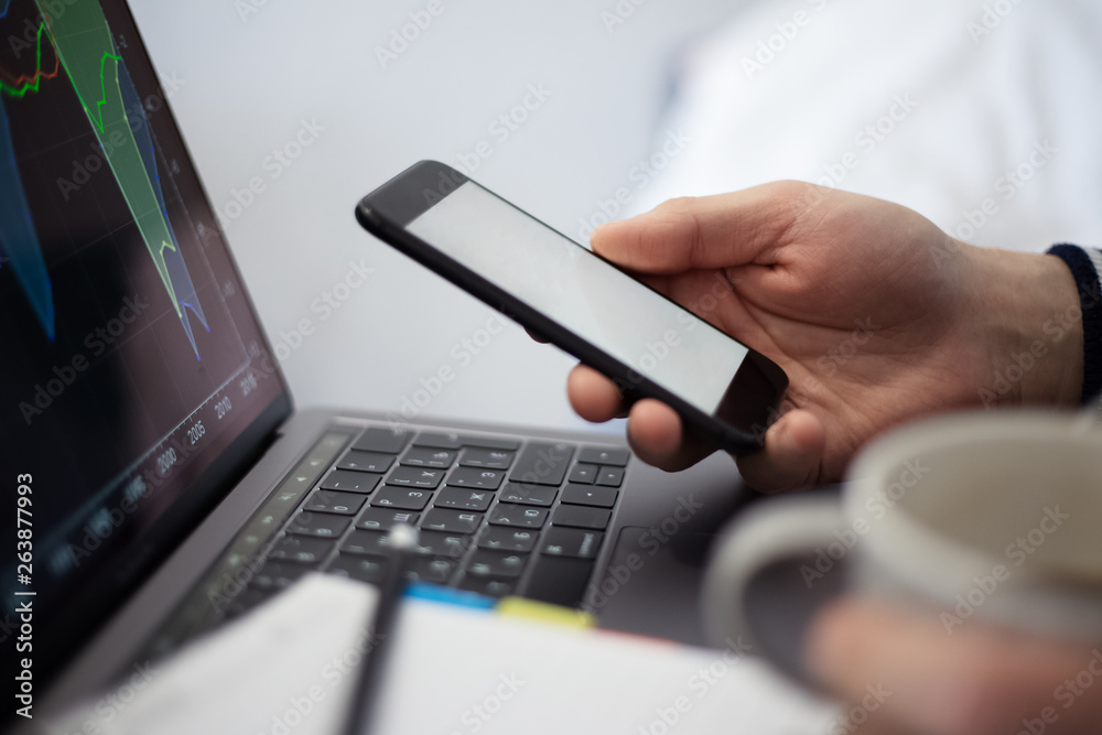 Close-up of businessman analysing data of finance statistic on laptop. Holding smartphone with mockup and cup of coffee. Close-up of businessman analysing data of finance statistic on laptop. Holding smartphone with mockup and cup of coffee.