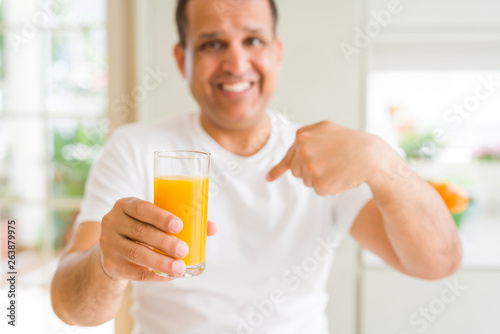 Middle age man drinking a glass of orange juice at home with surprise face pointing finger to himself