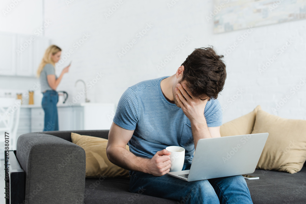 selective focus of upset man covering face while holding cup near laptop selective focus of upset man covering face while holding cup near laptop