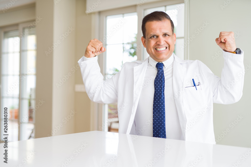 Middle age doctor man wearing medical coat at the clinic showing arms muscles smiling proud. Fitness concept.