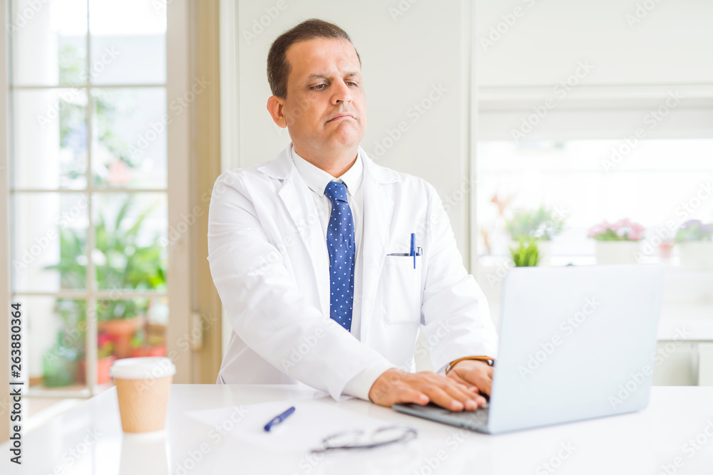 Middle age doctor man wearing white medical coat working with laptop at the clinic with serious expression on face. Simple and natural looking at the camera.