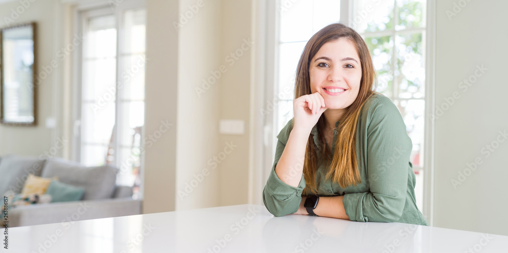 Beautiful young woman at home looking confident at the camera with smile with crossed arms and hand raised on chin. Thinking positive.