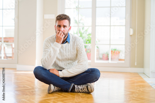 Handsome man wearing casual sweater sitting on the floor at home looking stressed and nervous with hands on mouth biting nails. Anxiety problem.