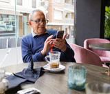 Handsome middle age senior man drinking coffee at restaurante, smiling happy enjoying and relaxing retirement using smartphone