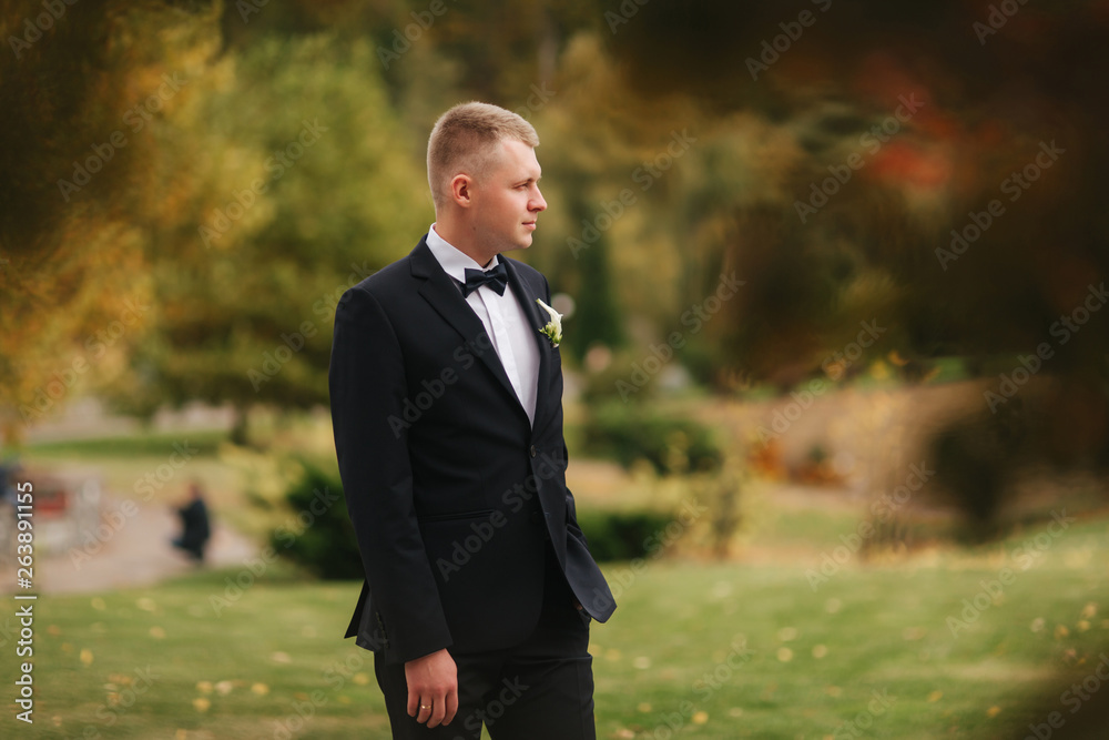 Handsome man in black suit standing outside in autumn. Groom waiting for bride Handsome man in black suit standing outside in autumn. Groom waiting for bride