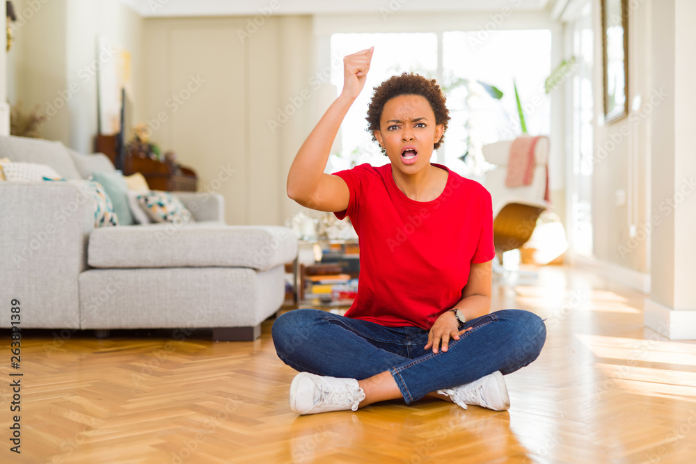 Young beautiful african american woman sitting on the floor at home angry and mad raising fist frustrated and furious while shouting with anger. Rage and aggressive concept. Young beautiful african american woman sitting on the floor at home angry and mad raising fist frustrated and furious while shouting with anger. Rage and aggressive concept.