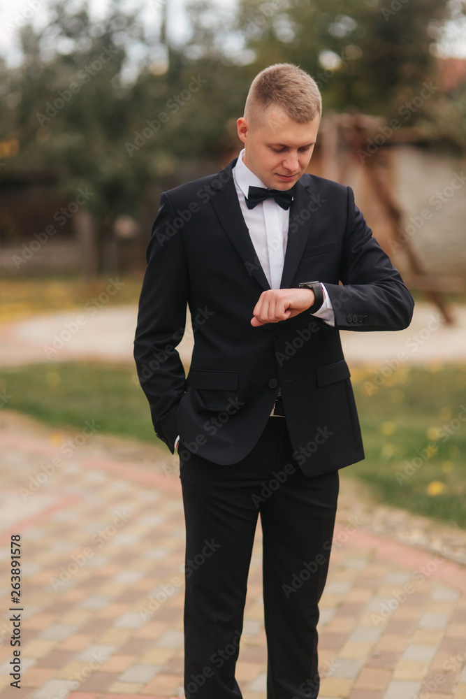 Handsome man in suit look at the watch outside, autumn. Man in black suit with beautiful tie bow Handsome man in suit look at the watch outside, autumn. Man in black suit with beautiful tie bow