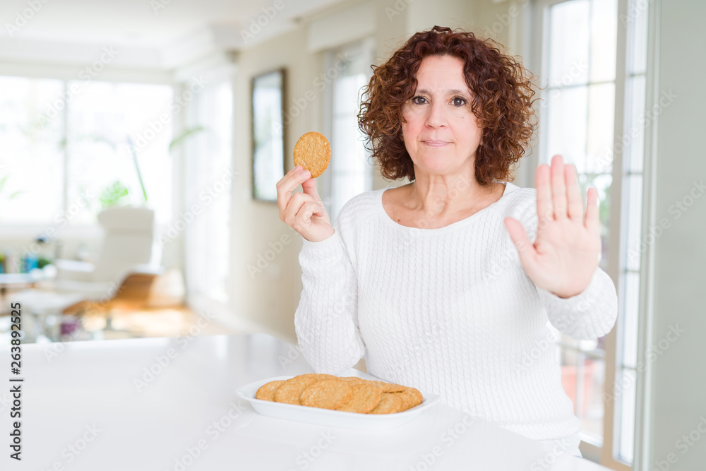 Senior woman eating healthy whole grain biscuit at home with open hand doing stop sign with serious and confident expression, defense gesture Senior woman eating healthy whole grain biscuit at home with open hand doing stop sign with serious and confident expression, defense gesture