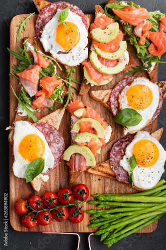 Mediterranean snacks set with brushettas, cheese variety, salami, eggs and salmon over black background on wooden board, top view