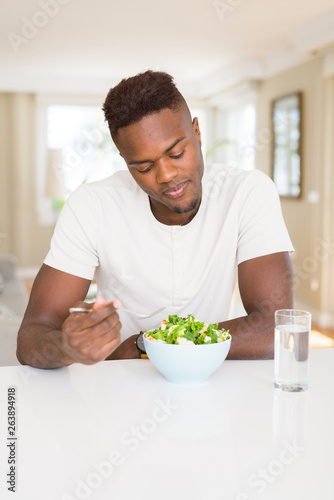 Handsome african young man eating a healthy vegetable salad using a fork to eat lettuce  happy and smiling sitting on the table