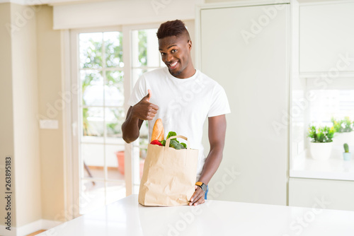 African american man holding paper bag full of fresh groceries happy with big smile doing ok sign, thumb up with fingers, excellent sign
