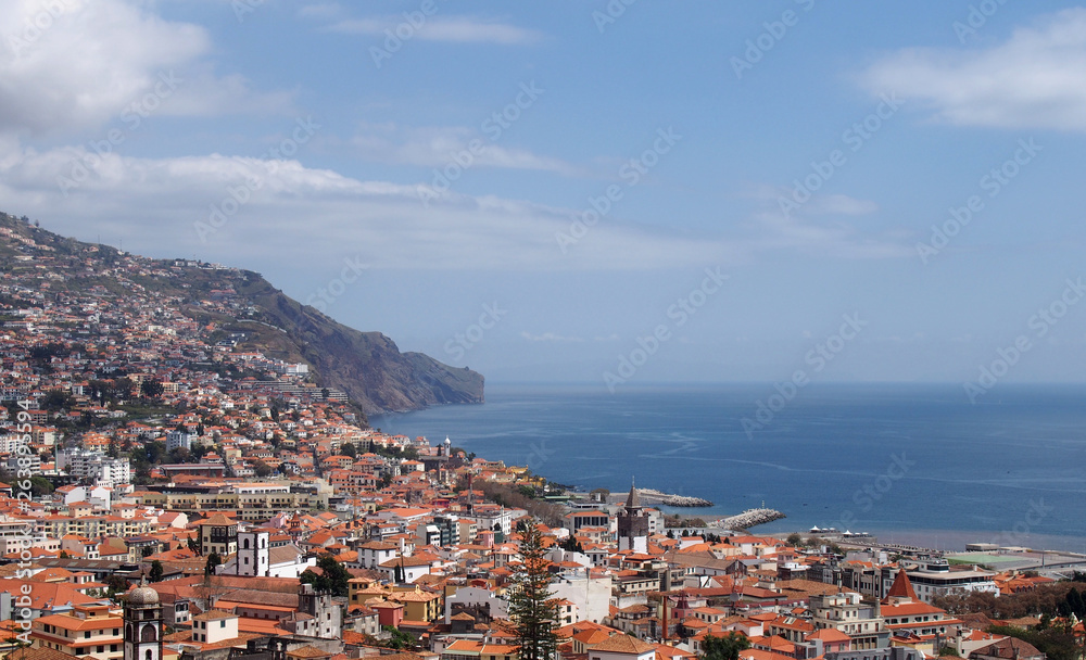 a sunlit view of the city of funchal from above with rooftops and buildings in front of a bright blue sea
