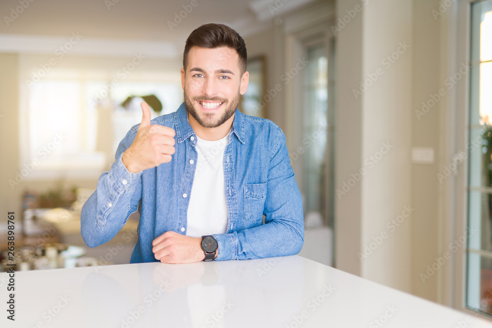 Young handsome man at home doing happy thumbs up gesture with hand. Approving expression looking at the camera with showing success.
