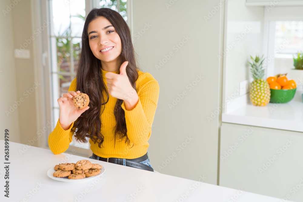 Young woman eating chocolate chips cookies at home happy with big smile doing ok sign, thumb up with fingers, excellent sign Young woman eating chocolate chips cookies at home happy with big smile doing ok sign, thumb up with fingers, excellent sign