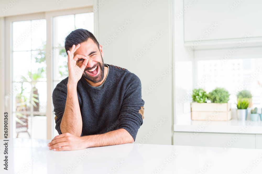 Handsome hispanic man wearing casual sweater at home doing ok gesture with hand smiling, eye looking through fingers with happy face.