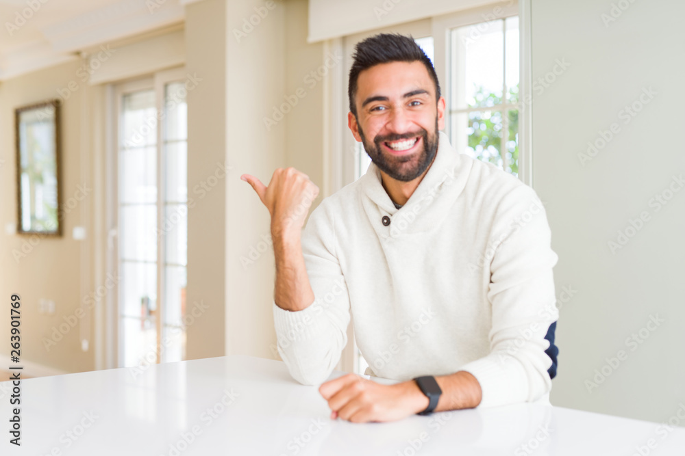 Handsome hispanic man wearing casual white sweater at home smiling with happy face looking and pointing to the side with thumb up.