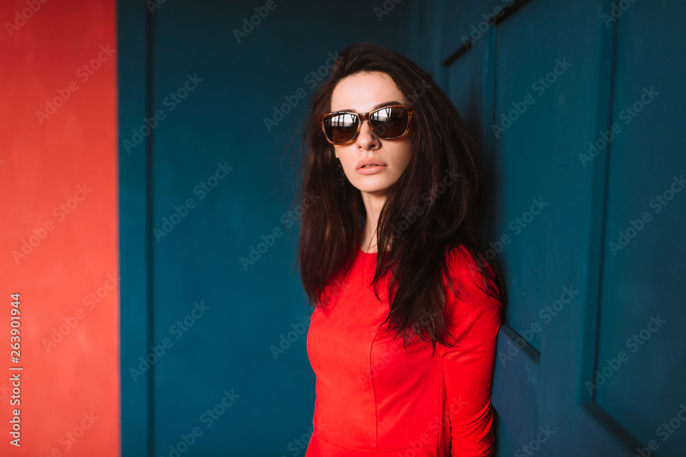 Beautiful fashion girl with dark long hair, spanish appearance in sunglasses and red elegant dress posing on blue red wall in studio. Indoor lifestyle portrait of stylish woman. Aamzing brunette babe. Beautiful fashion girl with dark long hair, spanish appearance in sunglasses and red elegant dress posing on blue red wall in studio. Indoor lifestyle portrait of stylish woman. Aamzing brunette babe.