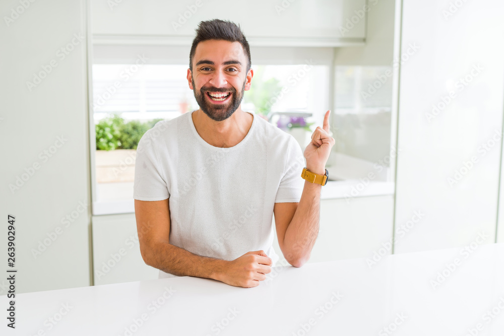Handsome hispanic man casual white t-shirt at home with a big smile on face, pointing with hand and finger to the side looking at the camera. Handsome hispanic man casual white t-shirt at home with a big smile on face, pointing with hand and finger to the side looking at the camera.