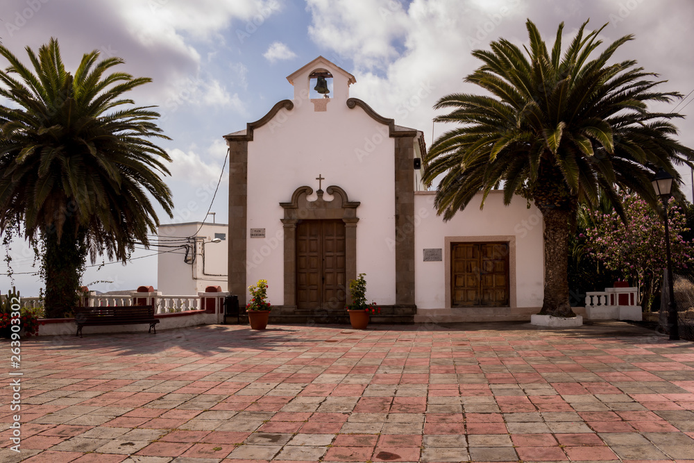 Obraz premium Church with a white facade. Heavy door and belfry with bells. Located on a small square. Cloudy sky. Plm trees beside the building. Arico Viejo, Tenerife, Canary Islands, Spain.