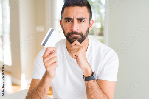 Handsome hispanic man holding credit card serious face thinking about question, very confused idea